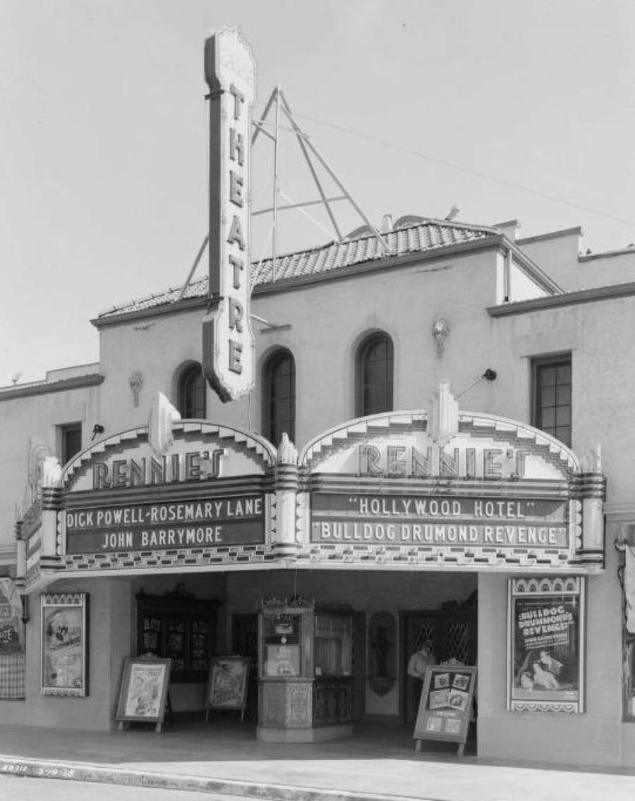 Black-and-white photo of an old movie theater with a large vertical "THEATRE" sign and marquee advertising “Hollywood Hotel” and “Bulldog Drummond’s Revenge.” Posters are displayed near the entrance.