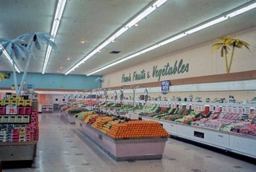 A grocery store produce section with neatly arranged fruits and vegetables, two decorative palm trees, and a sign reading "Fresh Fruits & Vegetables" on the back wall. The area is brightly lit with overhead lights.