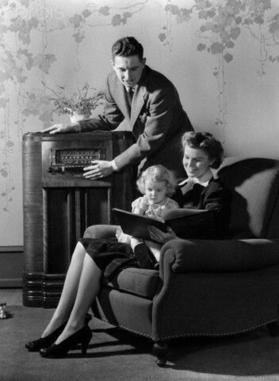 A man adjusts a vintage radio while a woman sits in an armchair reading to a young child on her lap. The scene is indoors with a decorated wall in the background.