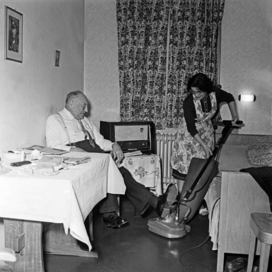 An elderly man sits at a dining table watching a woman vacuum the floor in a modest living room with patterned curtains, a radio, and a small couch.