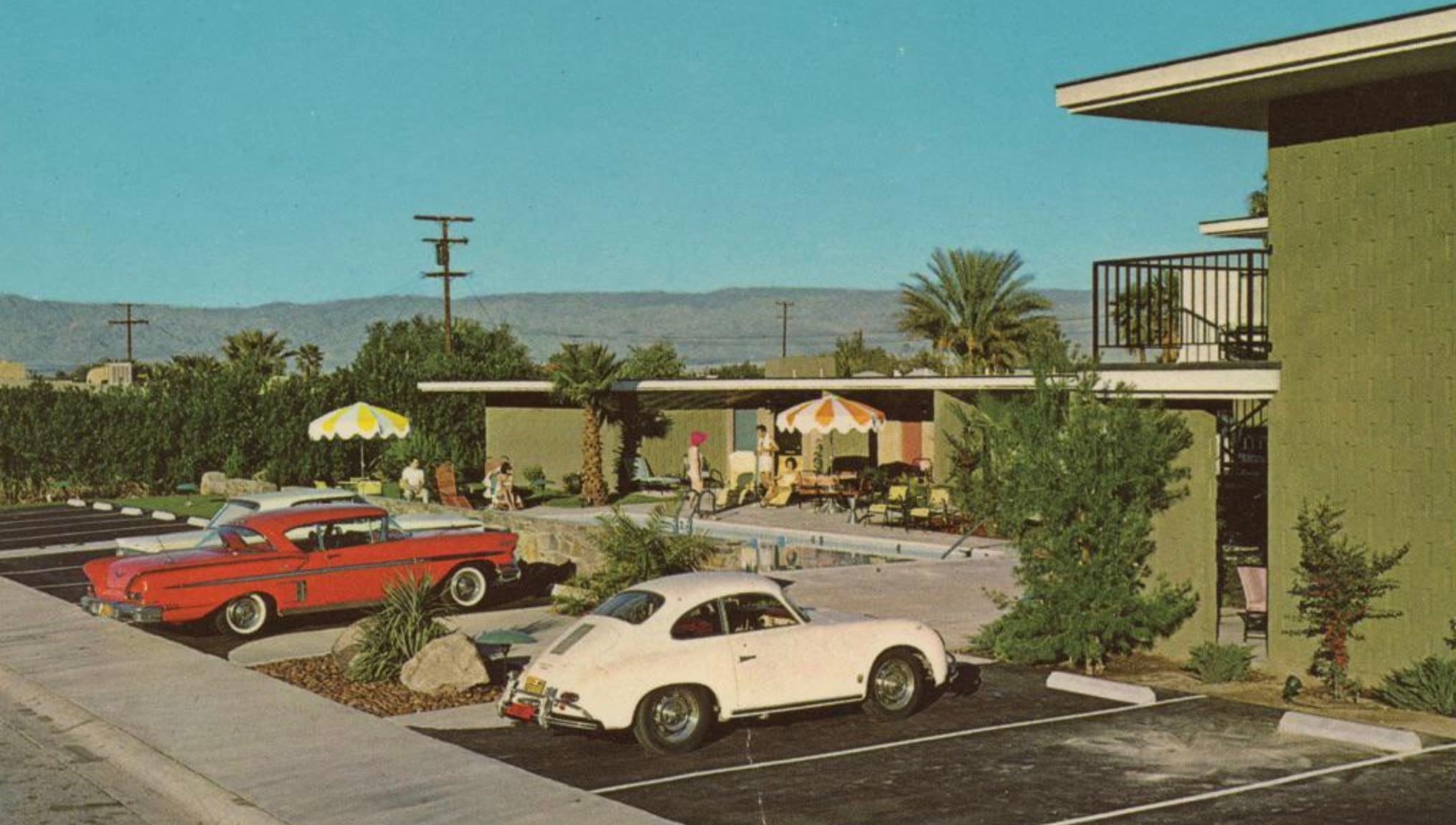 Vintage scene of a motel with a pool, people relaxing under yellow and orange umbrellas, a red classic car, and a white vintage car parked in front, with palm trees and mountains in the background.