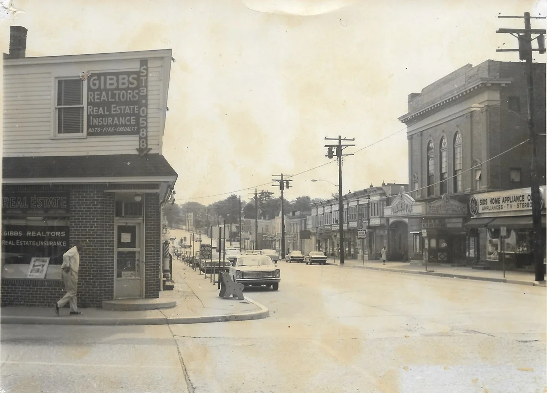 A vintage street scene shows an intersection with old cars, utility poles, and storefronts, including a Gibbs Realtors office and an appliance store. A person stands on the sidewalk near the corner. The image is in black and white.