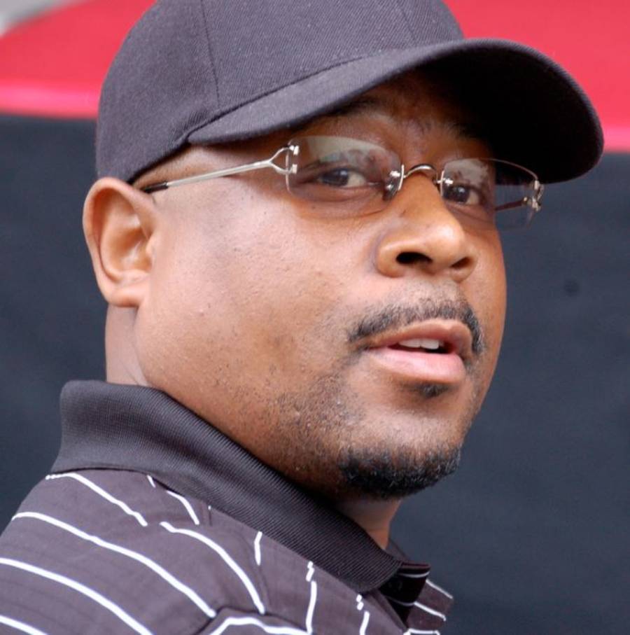 A man wearing a black cap, rimless glasses, and a black polo shirt with white stripes looks over his shoulder and smiles slightly. The background is blurred with shades of black and red.