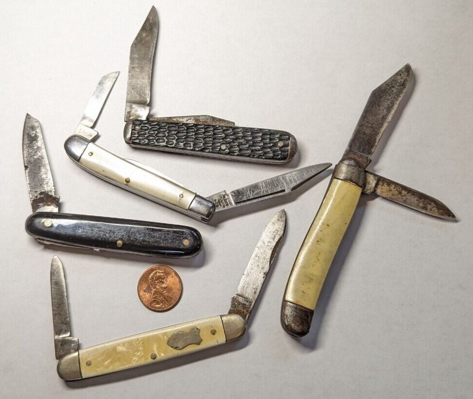 Five vintage pocket knives with worn blades and handles are arranged on a white surface, with a penny for size reference.