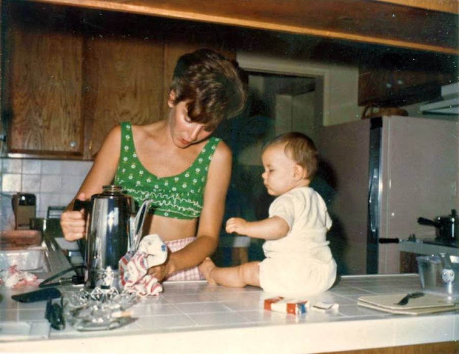 A woman in a green top washes dishes at a kitchen counter while a baby in white sits on the counter nearby, watching her.
