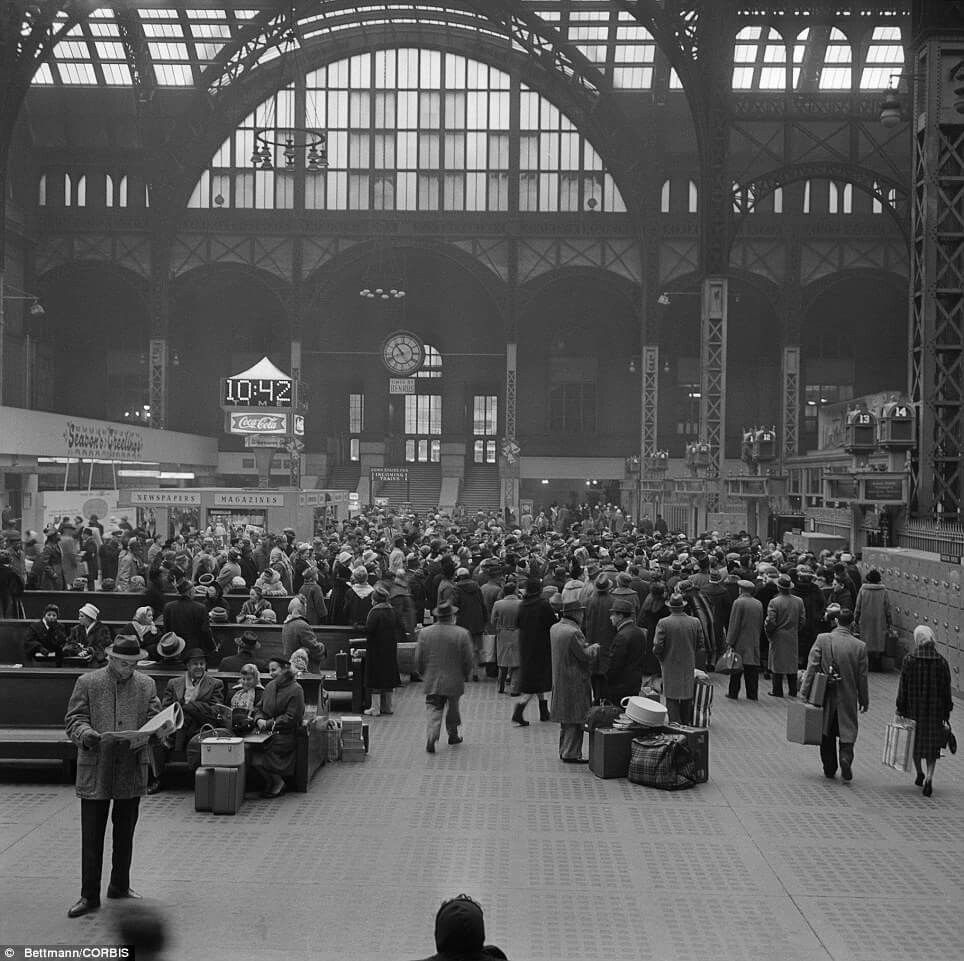 Black and white photo of a crowded train station with people waiting, some seated, others standing, beneath a large arched ceiling with a clock showing 10:42. Light streams in from tall windows above.