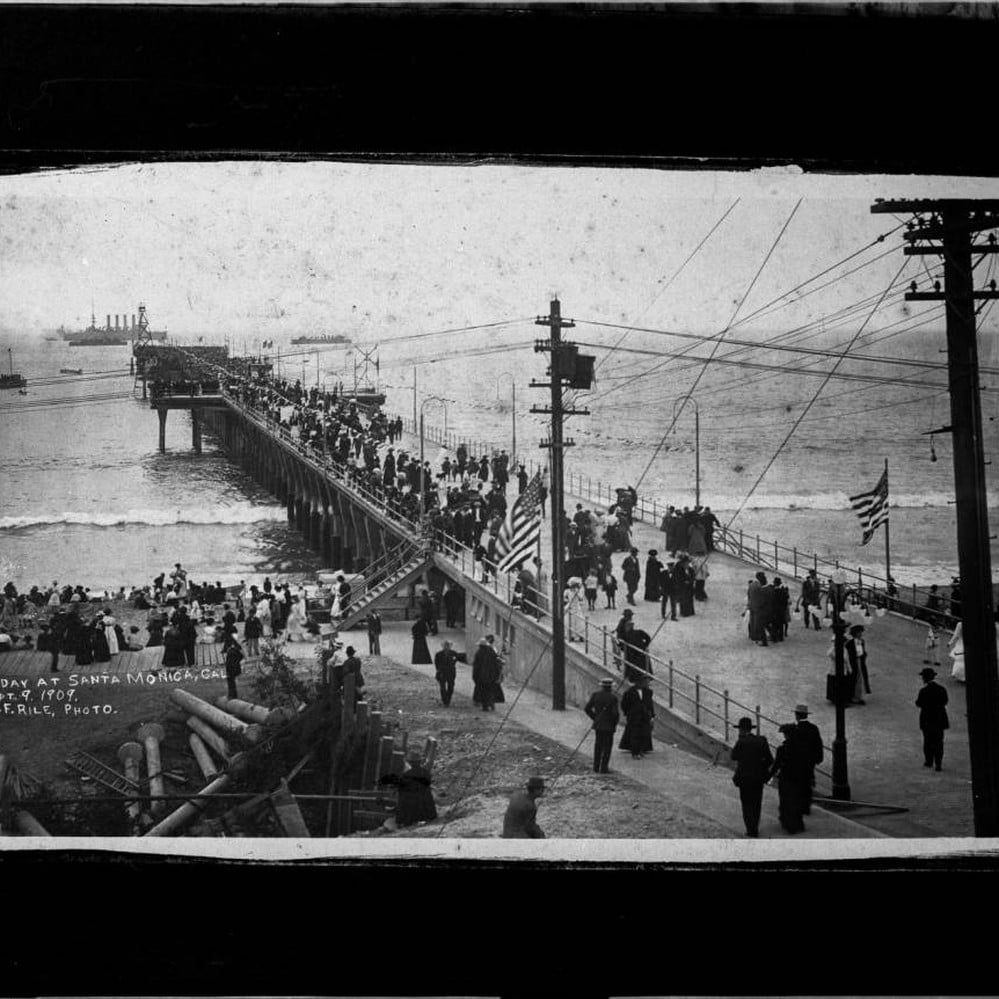 Black and white photo of a crowded pier in Santa Monica, California, with people walking along the pier and shoreline, American flags visible, and ships in the distance on the ocean.