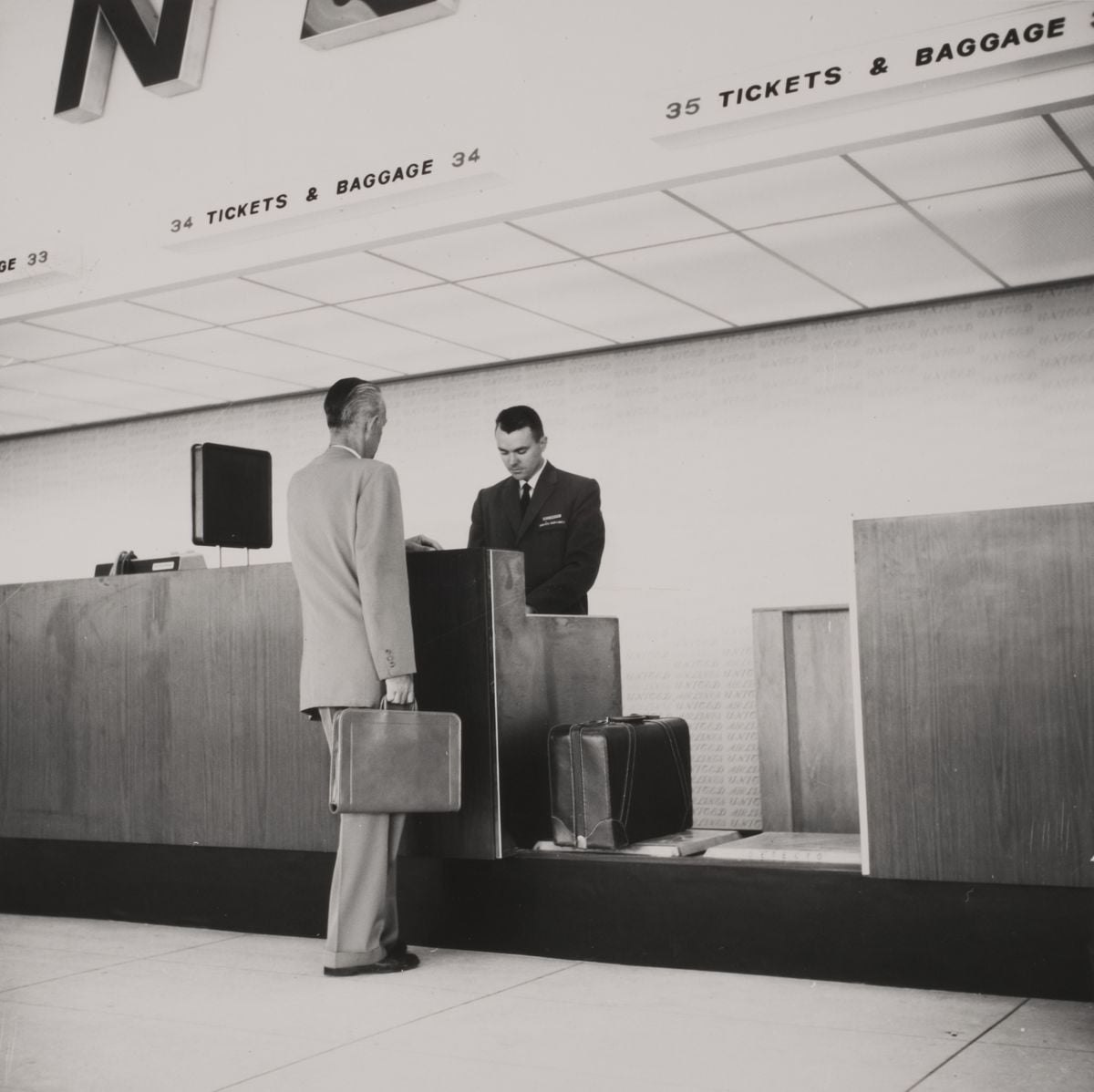A man in a suit holding a briefcase stands at an airline ticket and baggage counter, speaking with a uniformed attendant; two suitcases rest on the floor beside the counter.