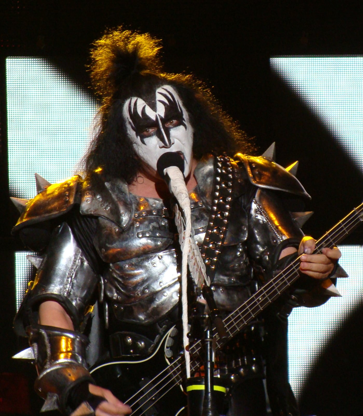 A musician in black and white face paint and spiked silver armor plays a guitar and sings into a microphone on stage.