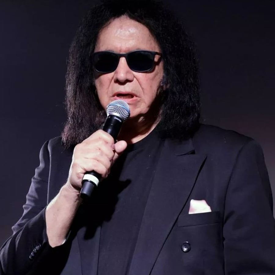 Man with long black hair wearing sunglasses and a black suit holds a microphone and speaks, standing against a dark background.