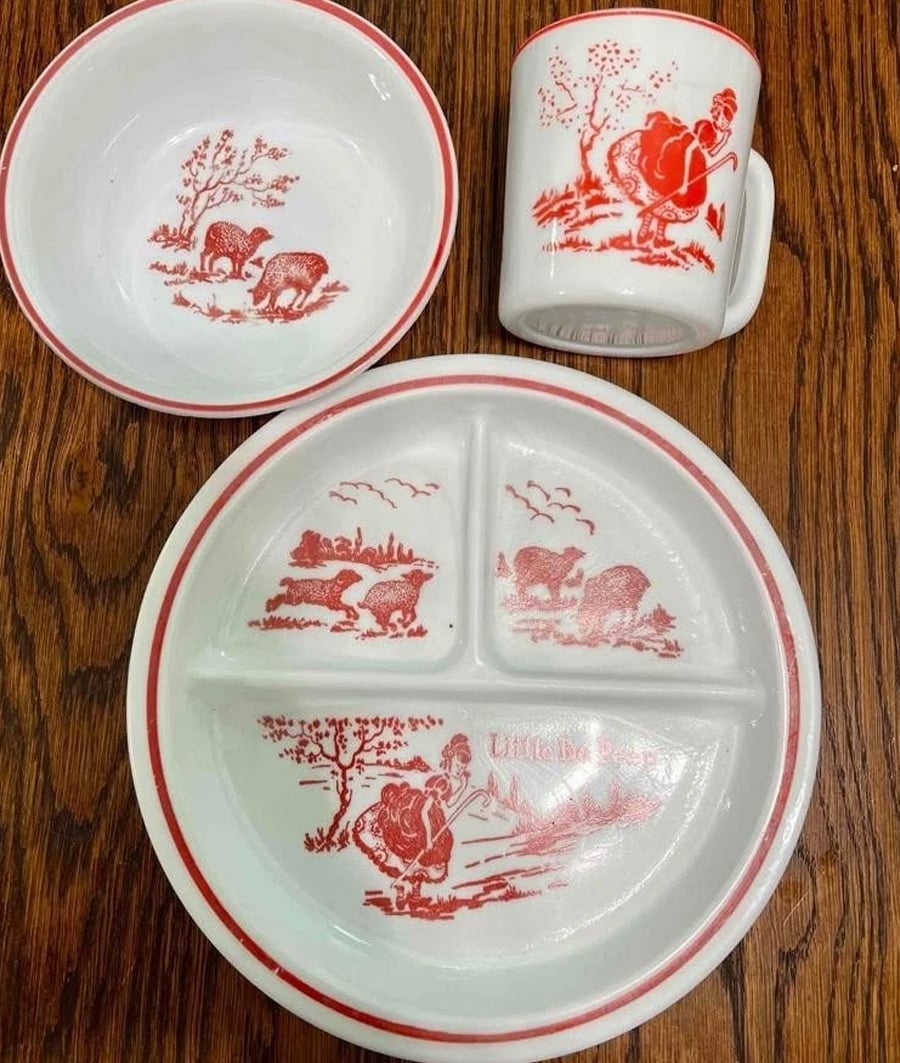 Children’s dish set with red "Little Bo Peep" illustrations on a plate, bowl, and mug, all decorated with red rim accents, displayed on a wooden surface.