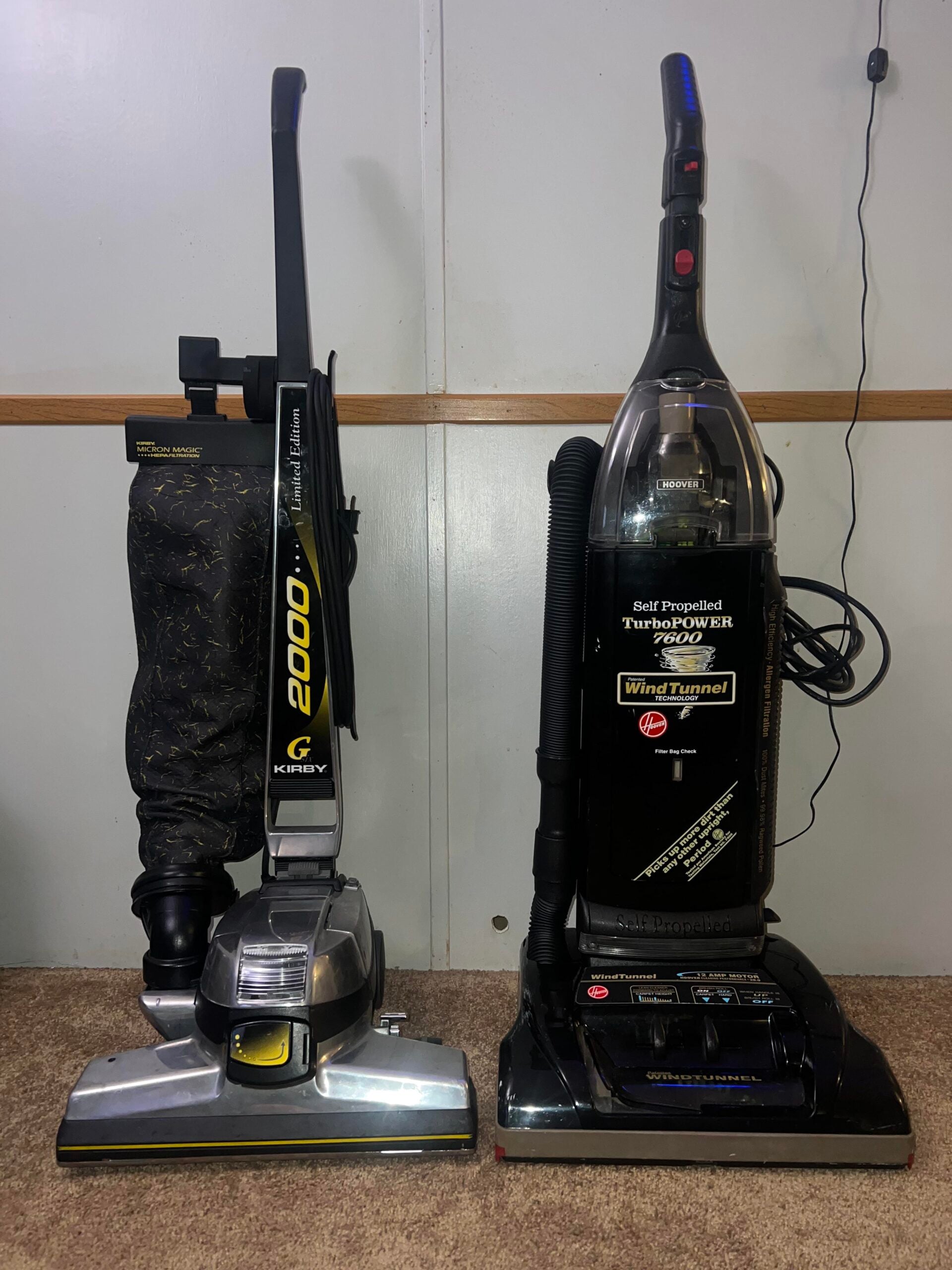 Two upright vacuum cleaners stand side by side on brown carpet against a plain wall. The left vacuum is silver with a black cloth bag; the right vacuum is black with a clear canister and visible labels on the front.