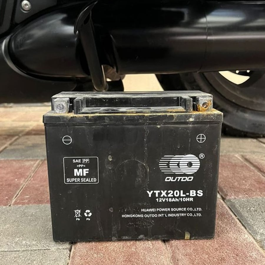 A black OUTDO YTX20L-BS motorcycle battery sits on a tiled surface with a vehicle part visible behind it. The battery displays technical details and manufacturer information on the front.