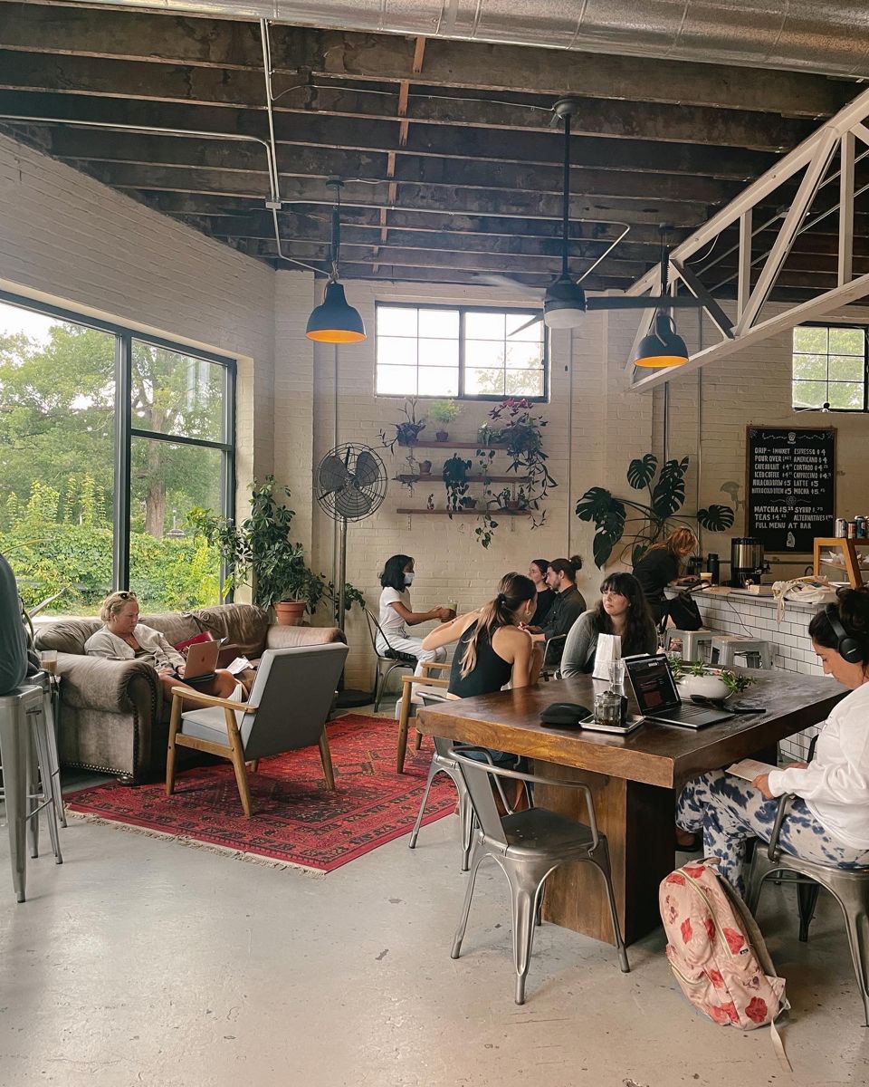 People work and socialize in a cozy, sunlit café with plants, large windows, and exposed ceilings. Some sit at tables with laptops, while others relax on sofas near a red rug. The atmosphere is casual and inviting.