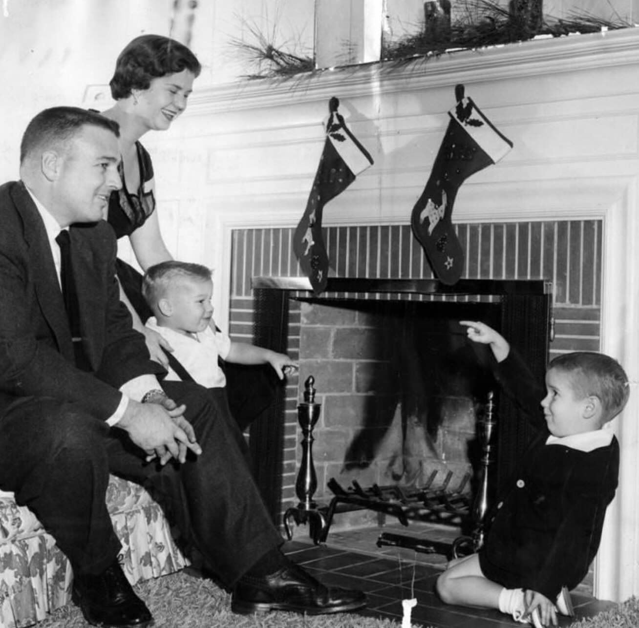 A family of four, dressed in formal attire, gathers in front of a fireplace with two Christmas stockings hanging above the mantle. One boy points toward the fire while the others smile and watch.