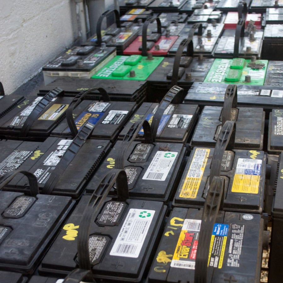 Several rows of used car batteries with various labels and colored caps are lined up against a wall, showing signs of wear and markings for recycling or disposal.