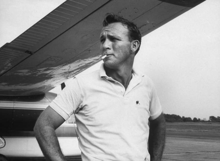 A man in a white polo shirt stands under an airplane wing, looking away with a cigarette in his mouth. The background shows an airstrip and trees.