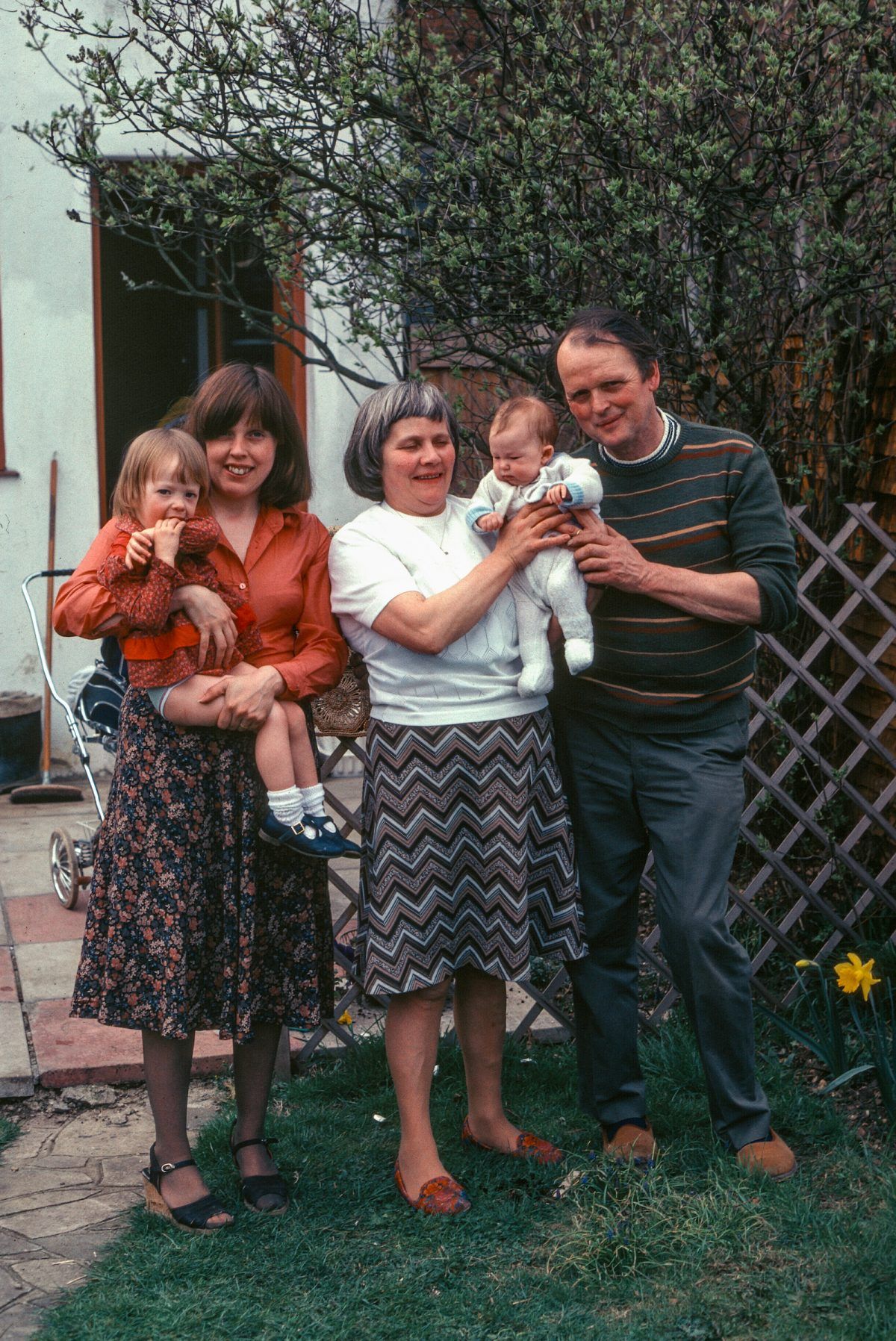 Four adults and two young children stand outside in a garden, smiling. One woman holds a toddler, and another woman holds a baby. The group is casually dressed, with some greenery and a wooden trellis in the background.