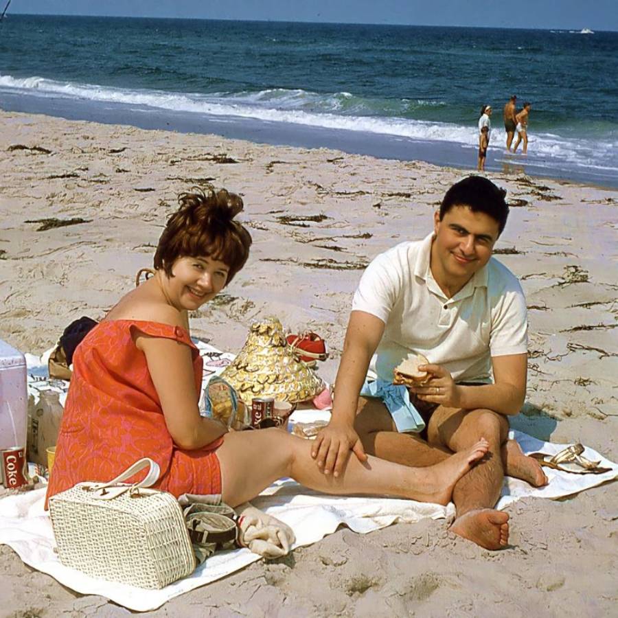 A woman and man sit on a blanket having a picnic at a sandy beach, smiling at the camera. The ocean is in the background with a few people standing near the water.