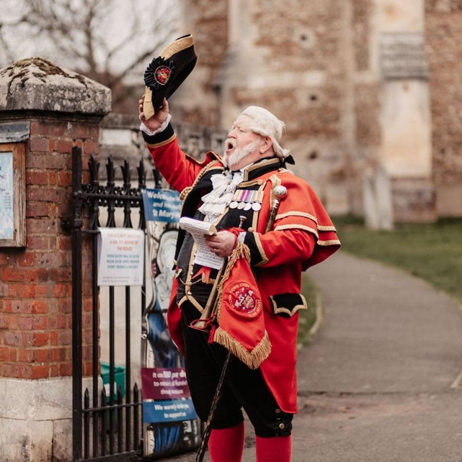 An older man dressed as a town crier in a red coat and black hat raises his hat and calls out near a brick wall and iron gate, holding papers in his other hand.