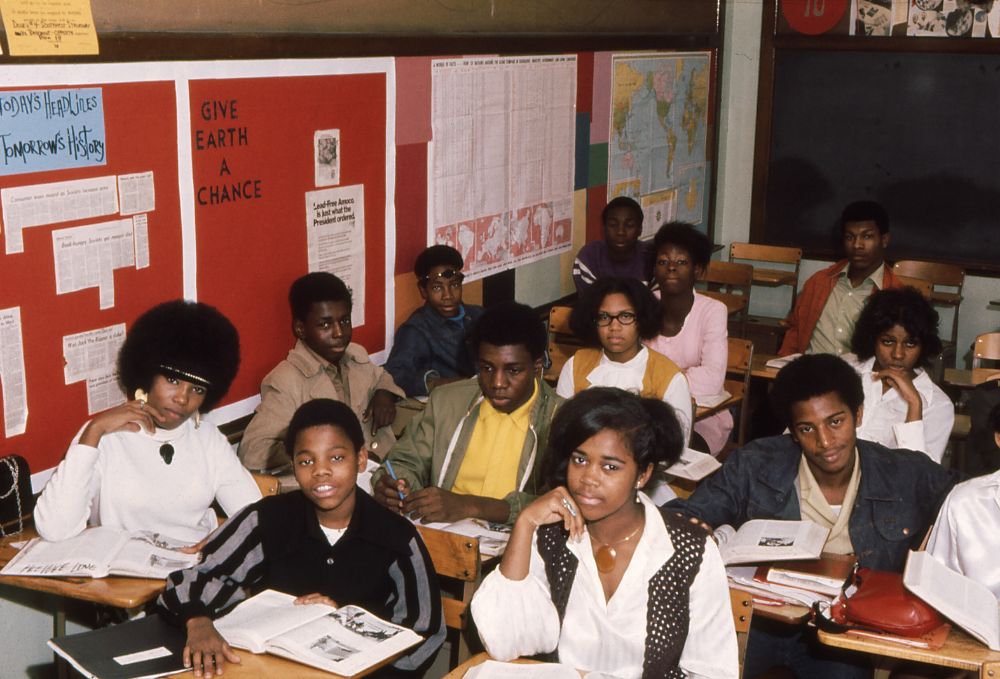 A classroom of Black students sit at desks with books open, looking toward the camera. Behind them are red bulletin boards with posters, maps, and the phrase "Give Earth a Chance.
