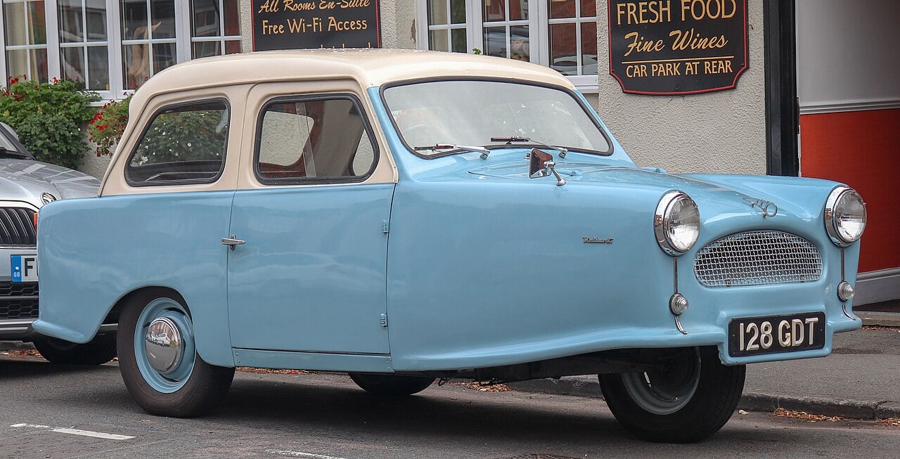 A light blue and cream vintage three-wheeled car parked on a street in front of a restaurant with signs in the background.