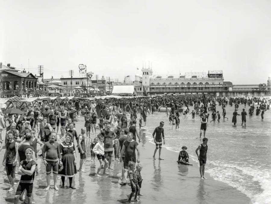 Black and white photo of a crowded beach with people in vintage swimwear, standing and wading in the water. Historic buildings and a pier are visible in the background under a clear sky.