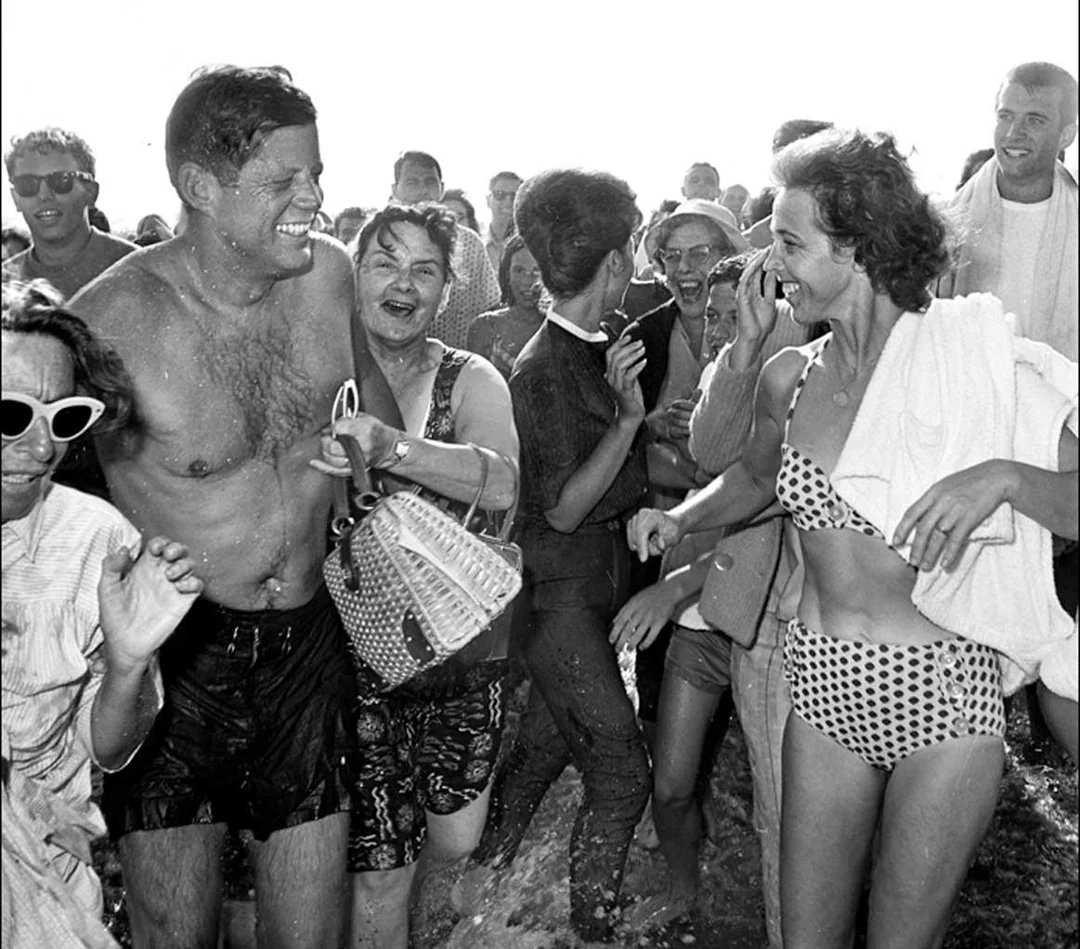 A joyful crowd surrounds a smiling man in swim trunks and a laughing woman in a polka-dot bikini on a bright, sunny beach.