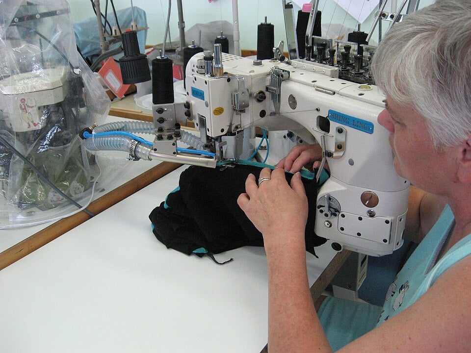 A person with short gray hair operates an industrial sewing machine, stitching black fabric with blue trim in a brightly lit workspace.
