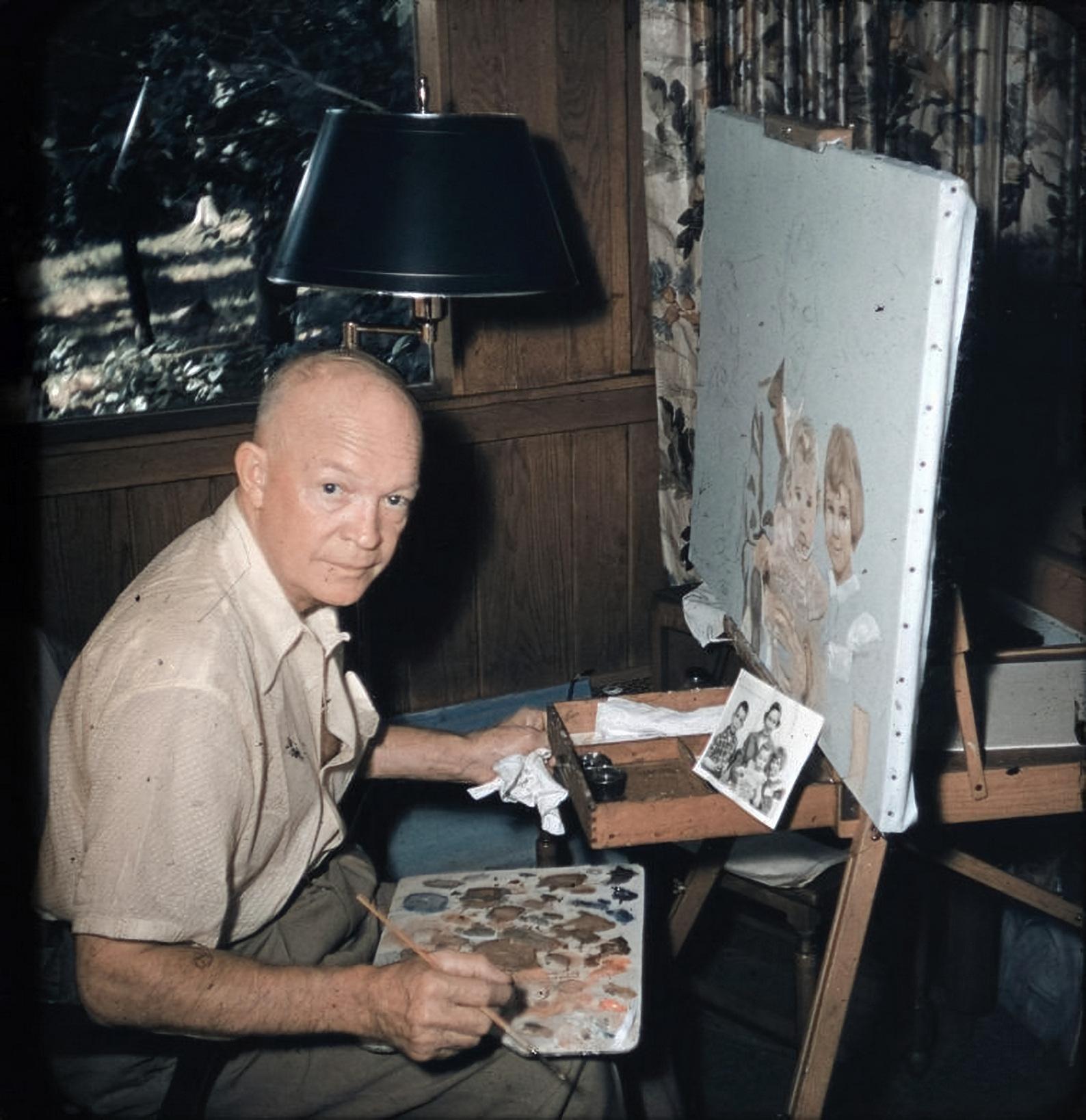 An older man sits at an easel, painting a portrait of a girl and a dog, with reference photos attached. He holds a paintbrush and palette, looking toward the camera in a cozy, wood-paneled room.
