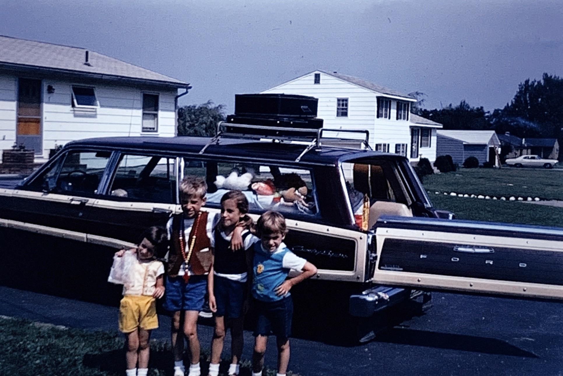 Four kids stand arm in arm in front of a packed Ford station wagon with tailgate open, in a suburban driveway on a sunny day. Two white houses and green lawns are visible in the background.