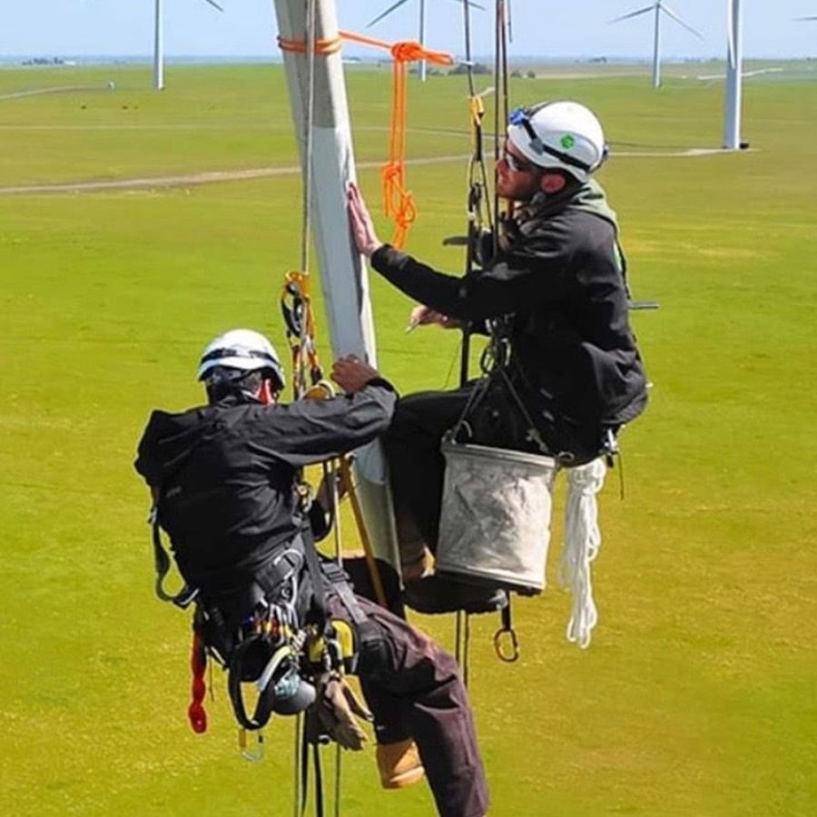 Two workers wearing safety harnesses and helmets perform maintenance while suspended on ropes from a wind turbine with grassy fields and other turbines in the background.