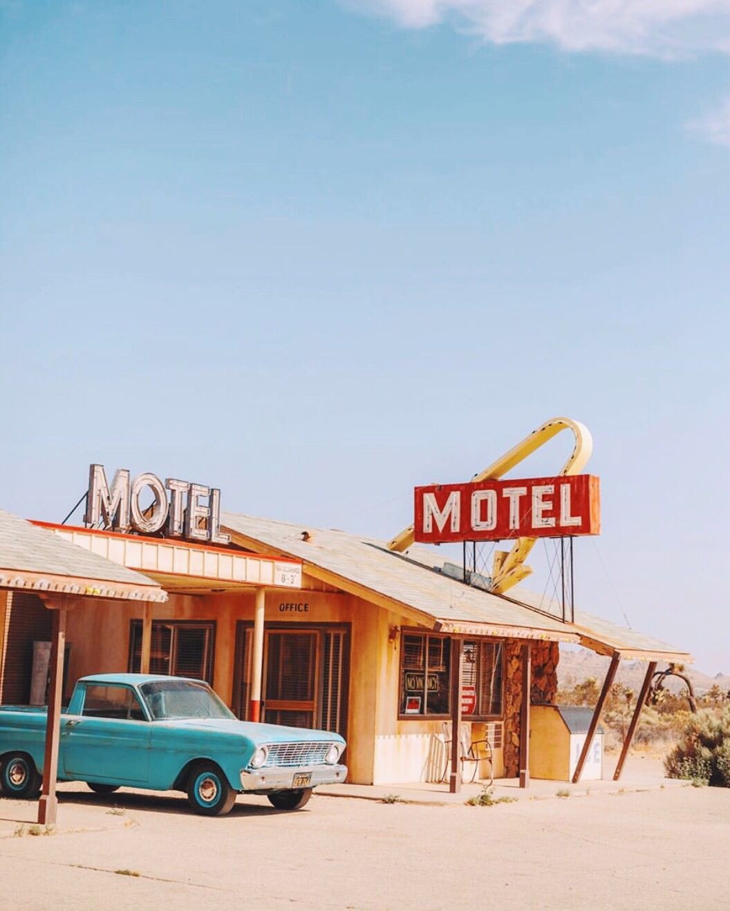A vintage turquoise car parked outside an old roadside motel with faded signage, under a clear blue sky in a desert setting.