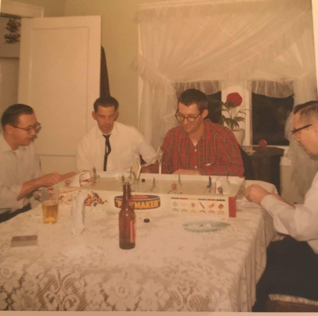 Four men sit around a table playing a tabletop hockey game. The table is covered with a white lace tablecloth, and there are drinks and game pieces on it. A window with sheer curtains and a red flower is in the background.