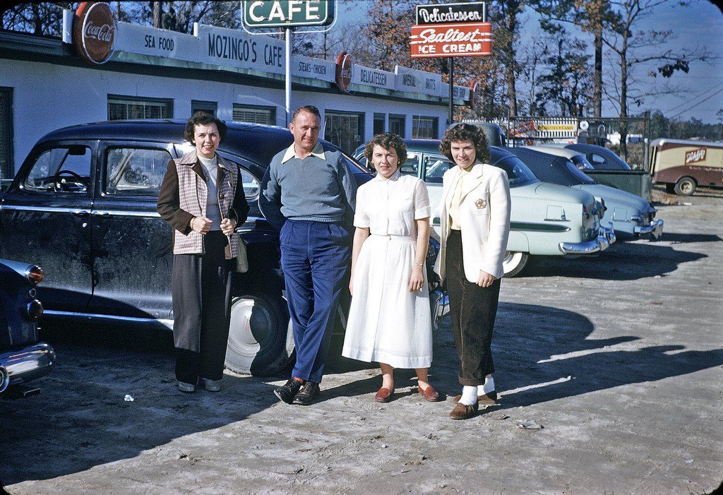 Four people stand in front of a vintage black car parked outside a diner called "Mozino's Café." Other classic cars are parked nearby, and a neon sign advertises Sealtest ice cream.