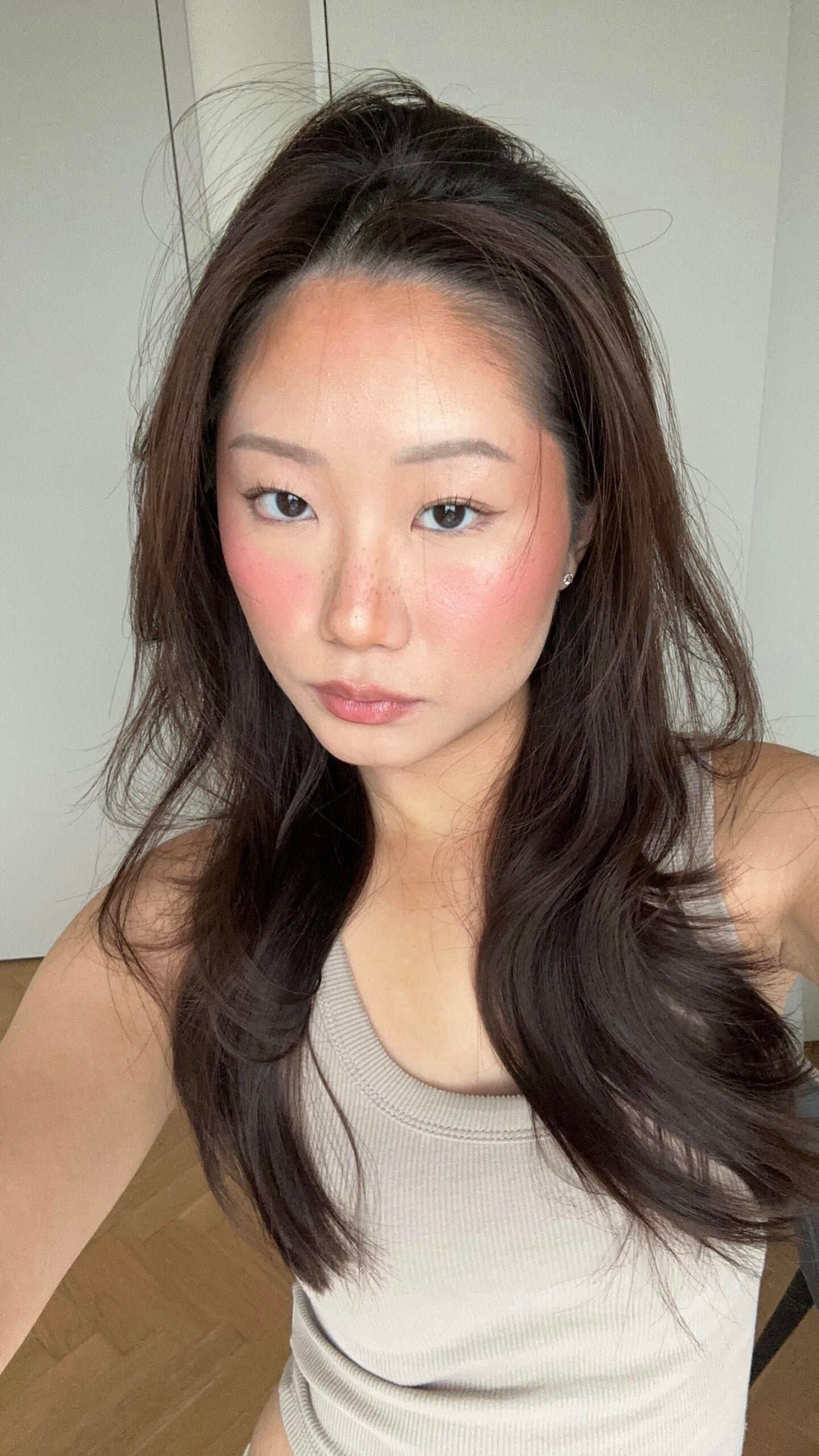 Woman with long dark hair wearing a beige tank top, posing for a selfie indoors with a neutral expression. She has rosy cheeks and natural makeup, and the background shows a white door and light-colored walls.
