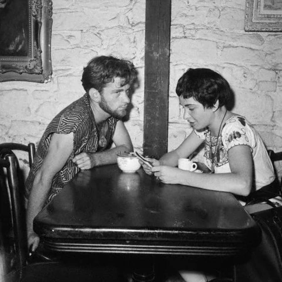 A man and woman sit across from each other at a cafe table. The woman looks at papers in her hand while the man leans in to listen. Both have drinks in front of them. Stone wall and framed picture are in the background.