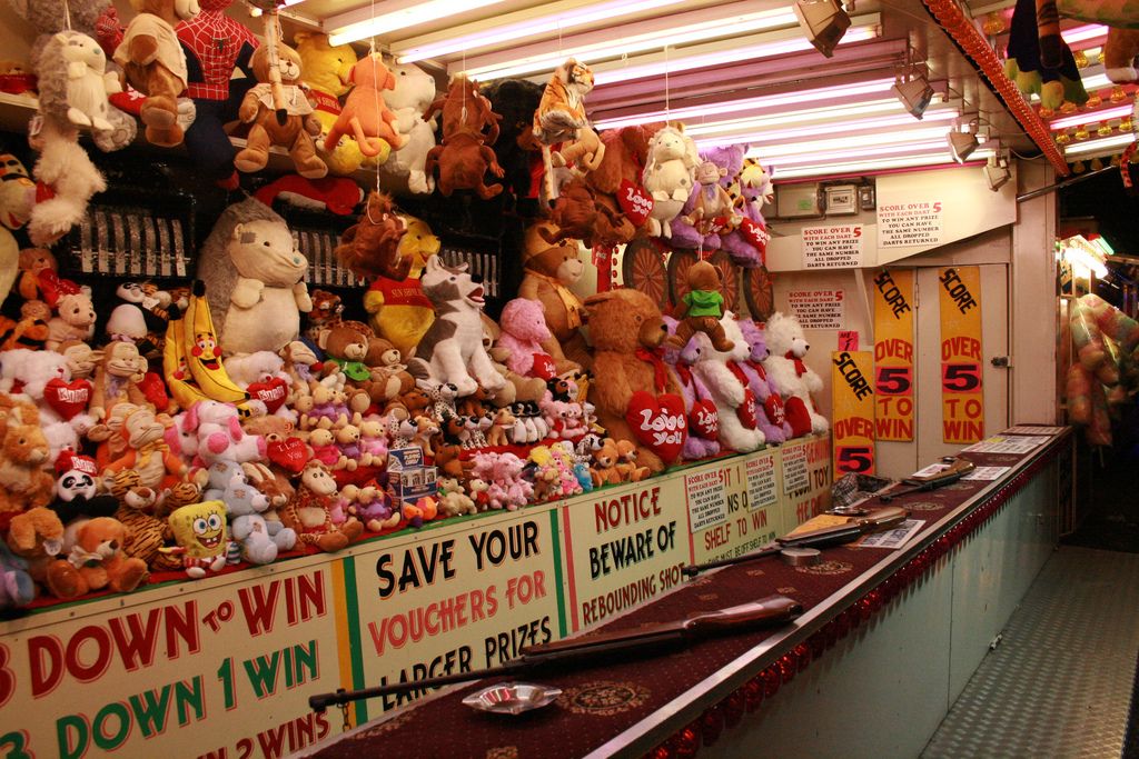 A carnival game booth with colorful stuffed animal prizes hanging on the wall and stacked on shelves, signs with game instructions, and metal trays with balls ready to be thrown.