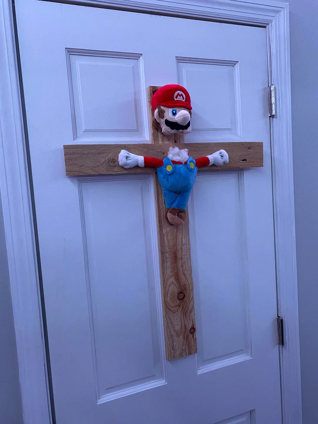 A Mario plush is attached to a wooden cross, mimicking a crucifix, and mounted on a white door. The lighting casts a slight blue tint over the scene.