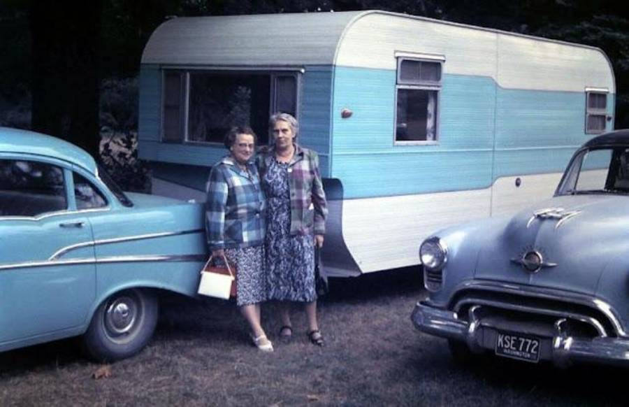 Two older women stand side by side in front of a blue and white camper trailer, flanked by two vintage blue cars. Trees and grass are visible in the background.