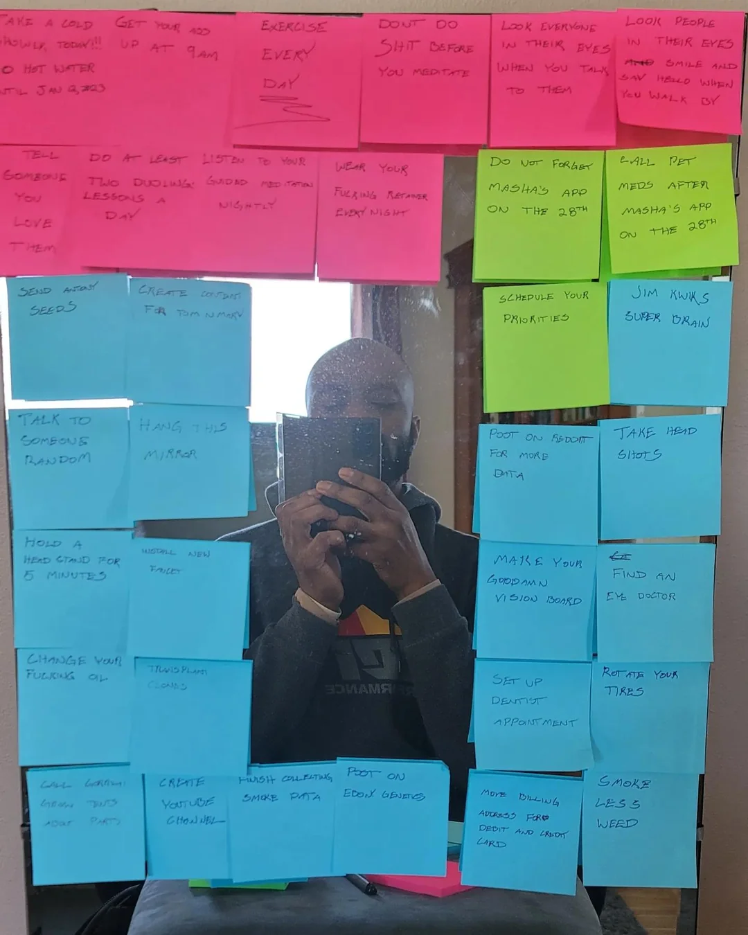 A mirror covered with colorful sticky notes containing handwritten reminders and tasks. A person is taking a photo of the mirror, and their reflection is visible in the center.
