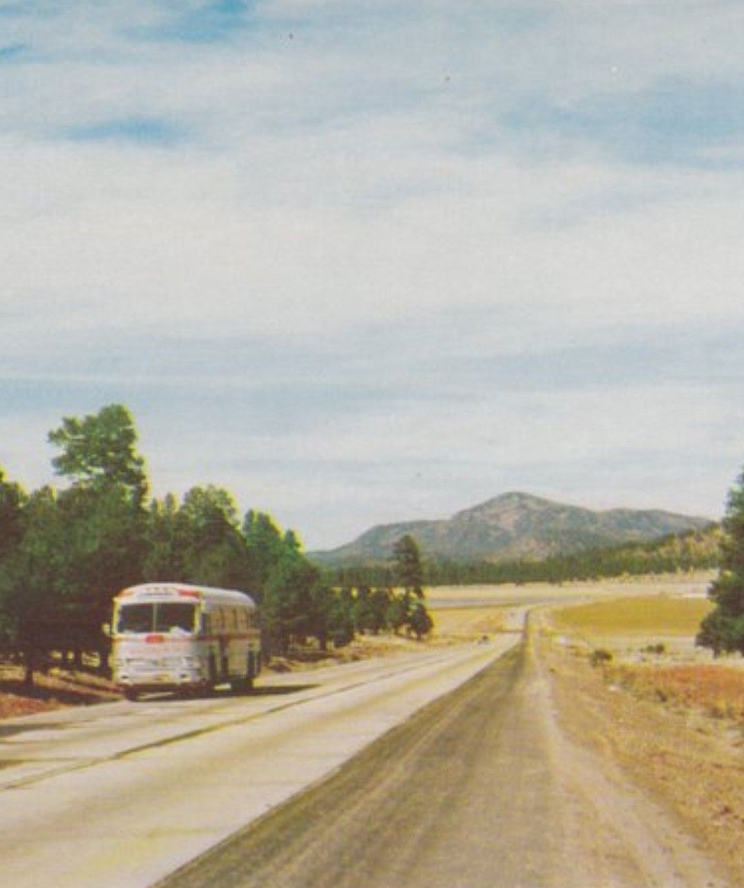 A vintage bus travels on a rural road lined with trees. The road stretches into the distance toward rolling hills and mountains under a partly cloudy sky.