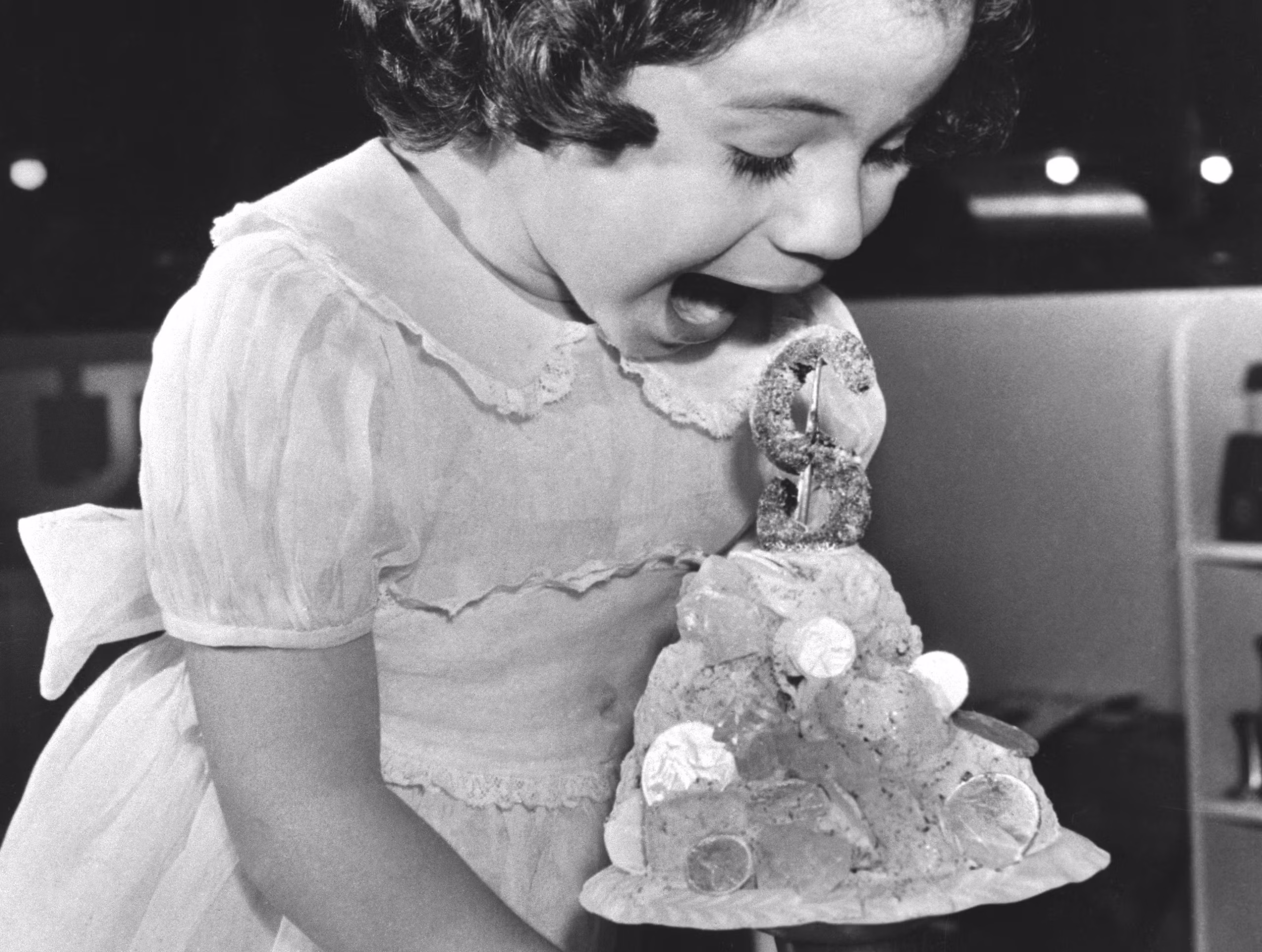 A young girl in a dress excitedly prepares to eat a large ice cream sundae topped with candy and a decorative figure.