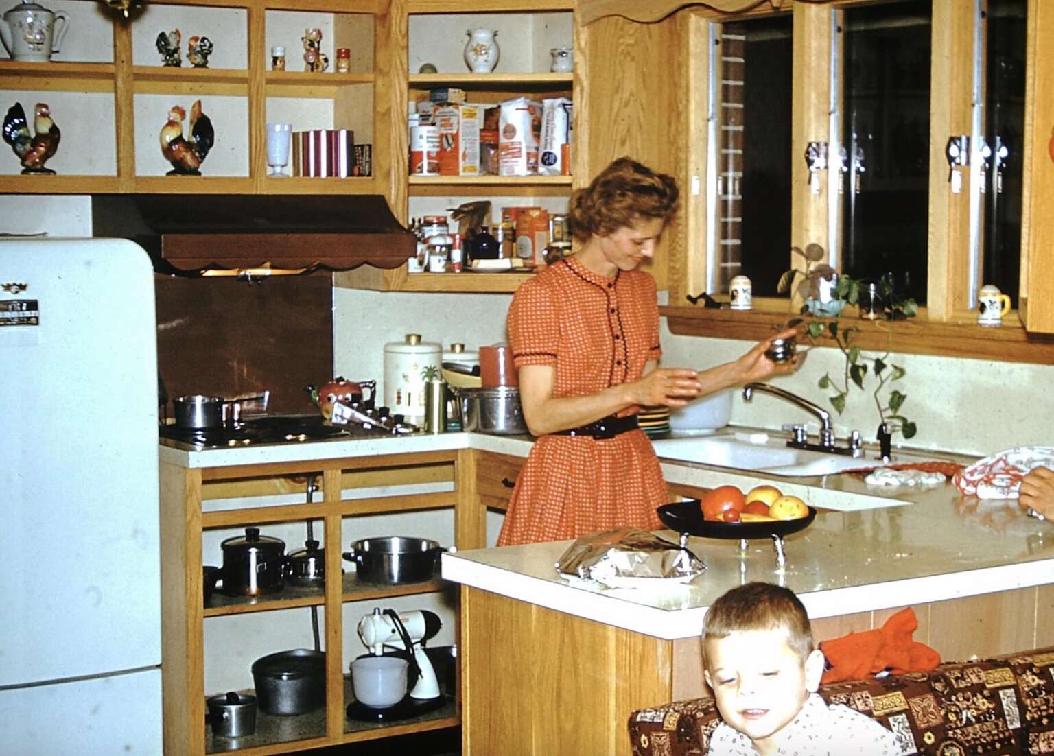 A woman in a red dress prepares food at a kitchen counter near a sink, while a young child sits on a chair in the foreground. The kitchen has wooden cabinets and various items on the shelves and counters.