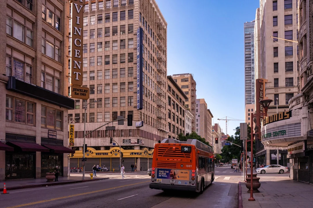 A city street lined with tall buildings, vintage signs for jewelry stores, and a Metro bus driving on the road in downtown Los Angeles on a clear, sunny day.