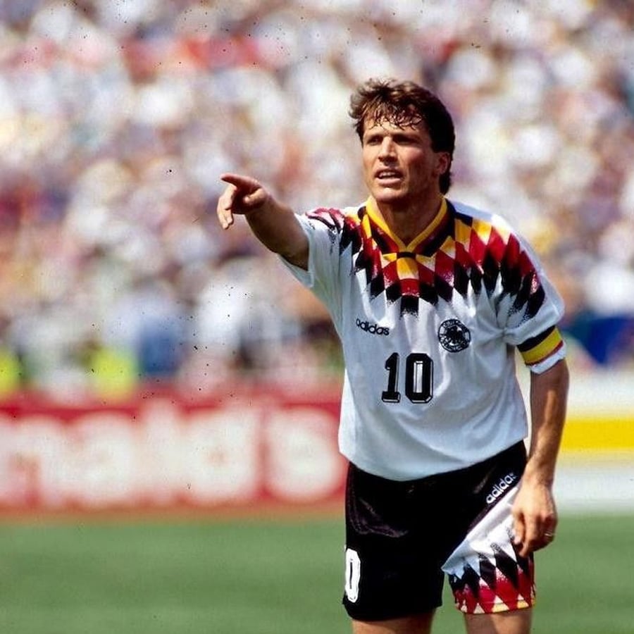 A soccer player in a white, black, and red Germany jersey with number 10 points forward on a sunny field during a match.