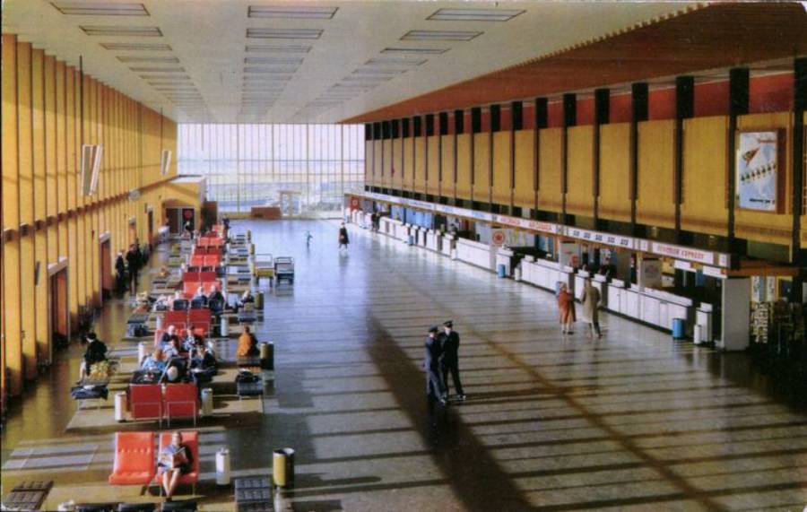 A spacious, retro-style airport terminal with large windows, yellow walls, red seats, and people sitting or walking near check-in counters. Sunlight streams in, casting long shadows on the shiny floor.