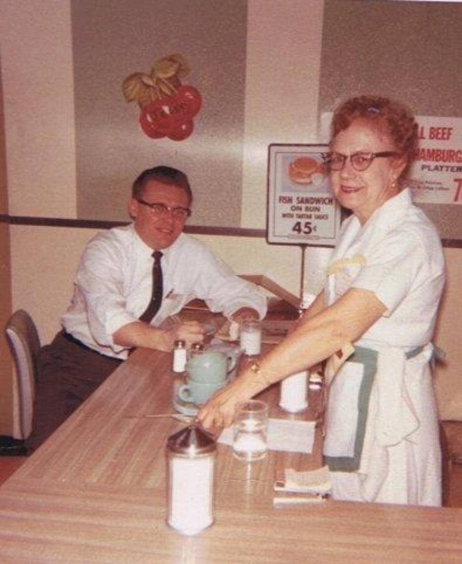 A man in a white shirt sits at a diner counter, smiling at an older woman in a waitress uniform. Both are by a table with coffee, sugar, and salt shakers. A sign advertises a 45-cent fish sandwich.