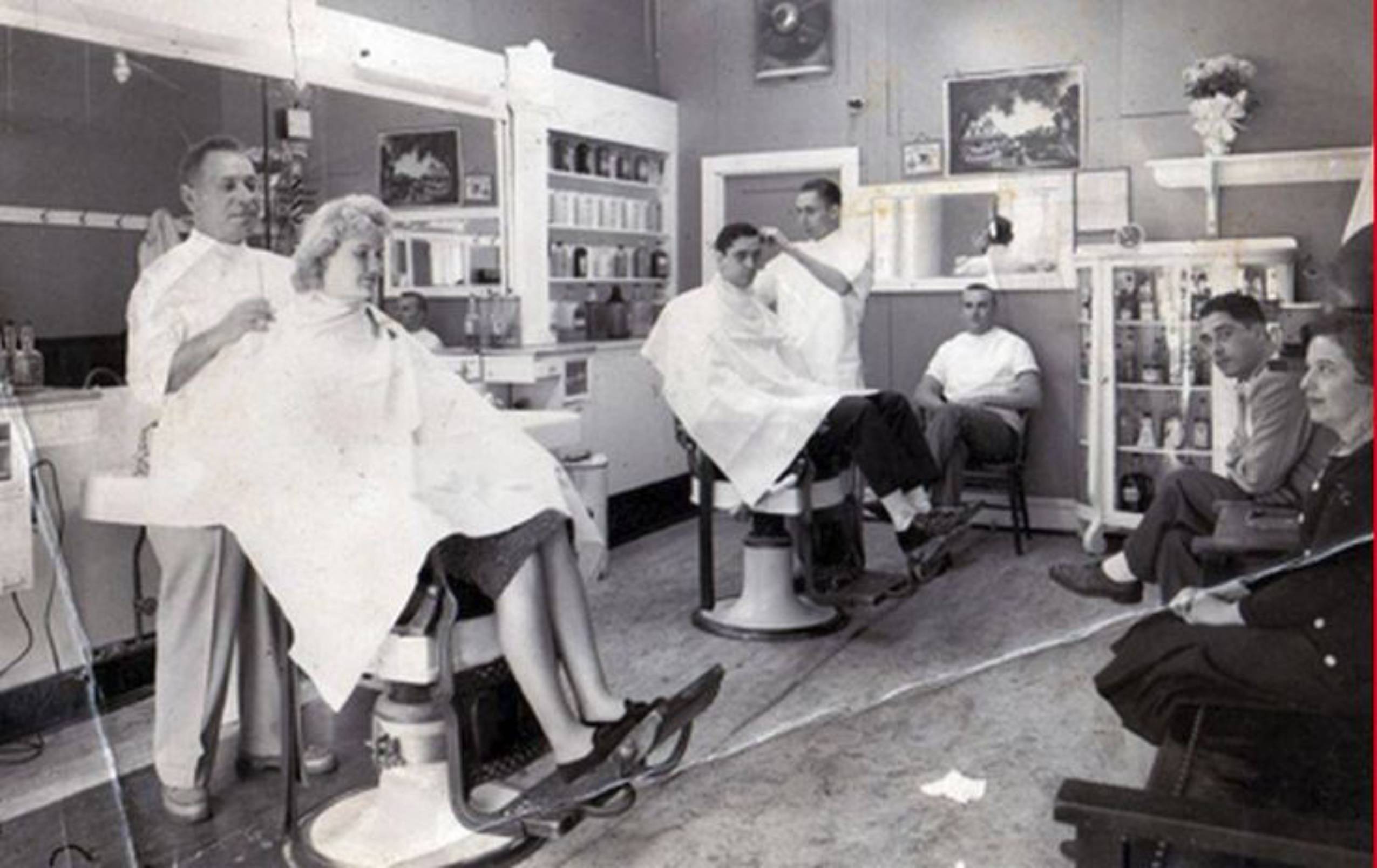 Black and white photo of a vintage barbershop with two clients getting haircuts, barbers working, and several people sitting and watching. Shelves with products and framed pictures decorate the walls.