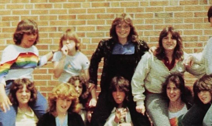 A group of teens poses in front of a brick wall. Some are sitting on others' shoulders. They wear casual 1980s-style clothing, including sweatshirts and a rainbow-striped shirt. Most are smiling or making playful faces.